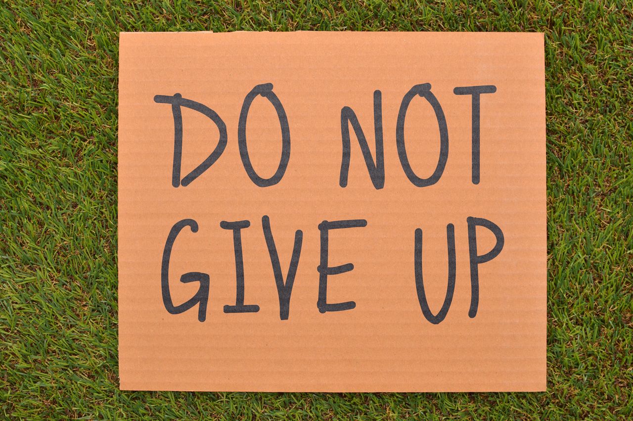 Cardboard sign reading 'Do Not Give Up' placed on green turf, symbolizing perseverance.